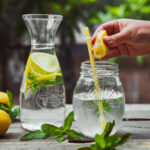 Hand squeezing lemon into a glass jar with water side view on wooden and yard background