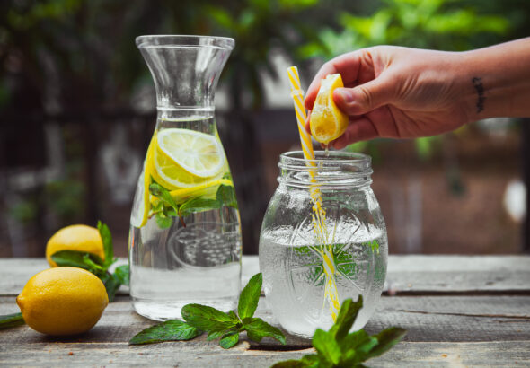 Hand squeezing lemon into a glass jar with water side view on wooden and yard background