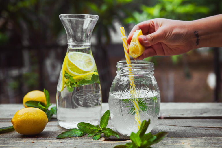 Hand squeezing lemon into a glass jar with water side view on wooden and yard background